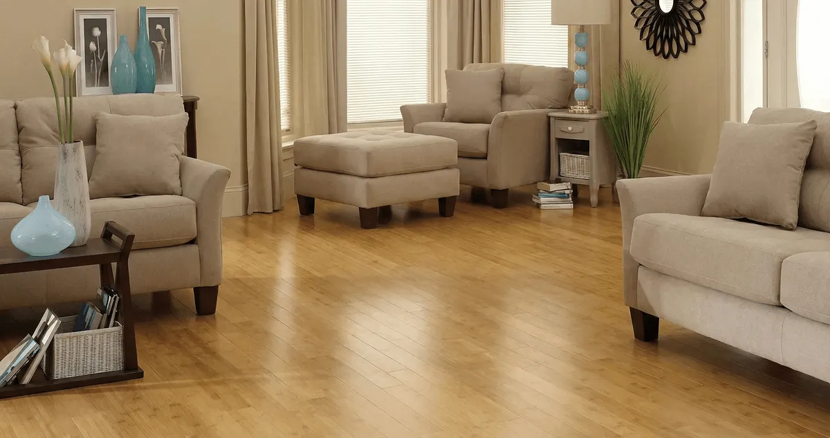 A living room with professionally installed bamboo flooring, beige sofas, a matching ottoman, a side table with a blue lamp, vases, framed pictures, and large windows with blinds and curtains in Chattanooga, TN